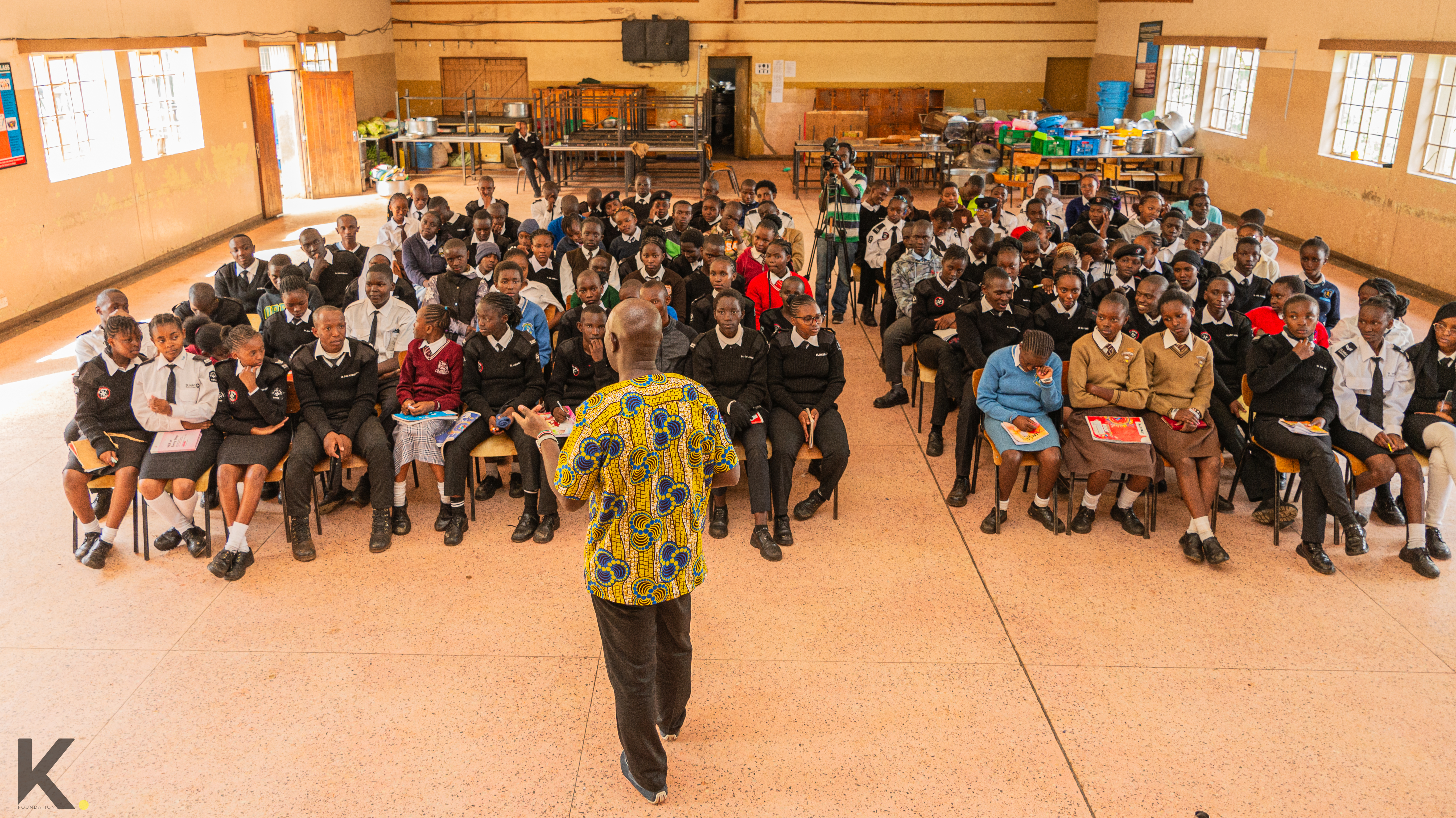 A large group of students in uniform seated in a classroom, listening to a teacher.