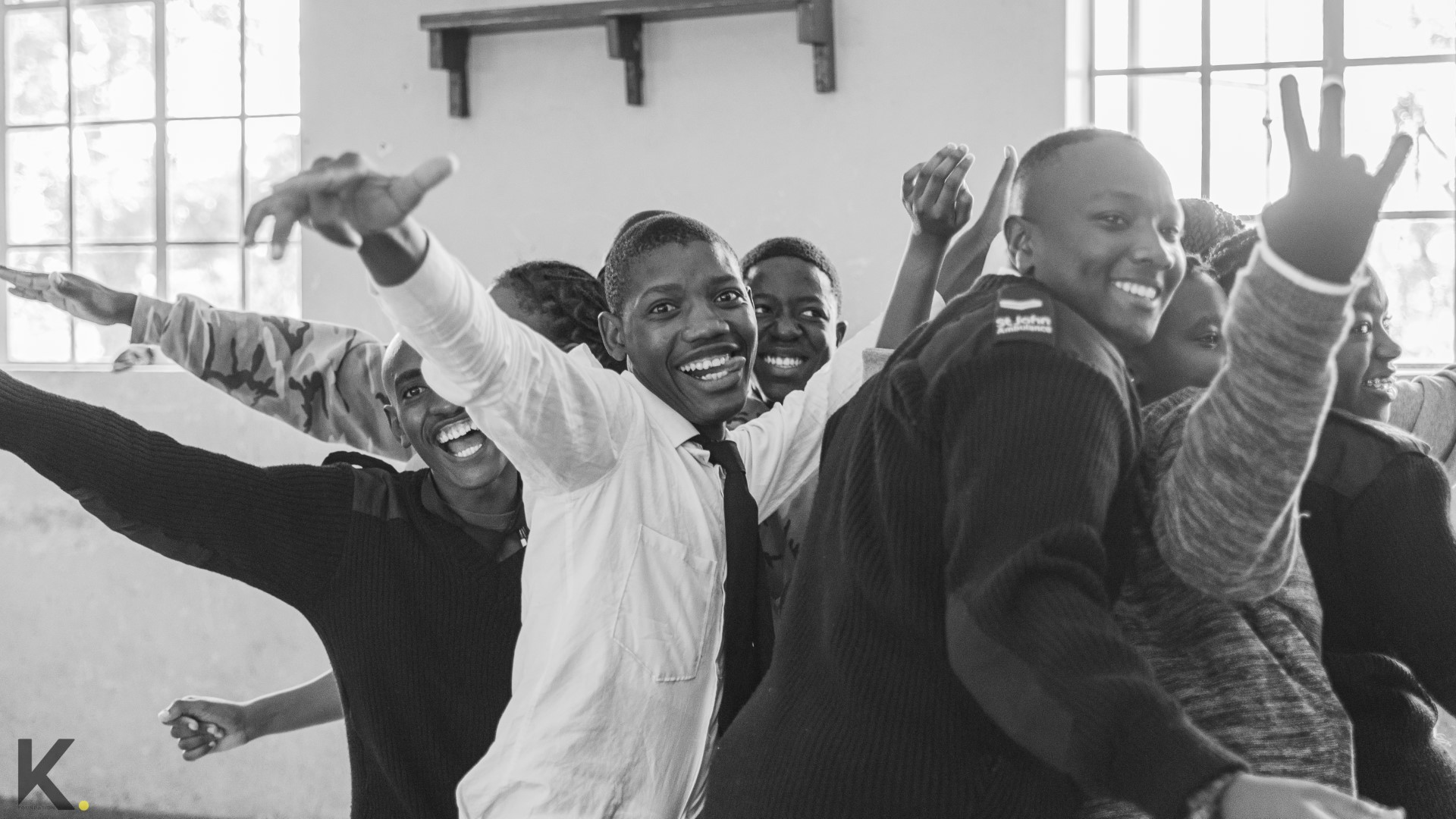 A group of smiling young people with raised hands, celebrating in a classroom.