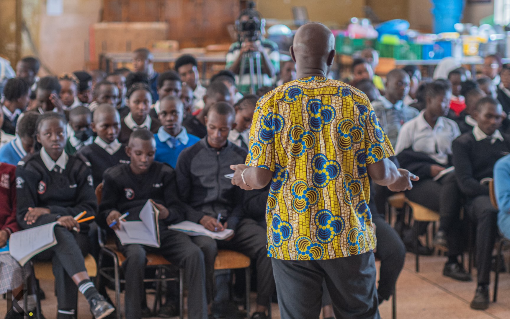 A teacher in a patterned shirt standing in front of a class of seated students.