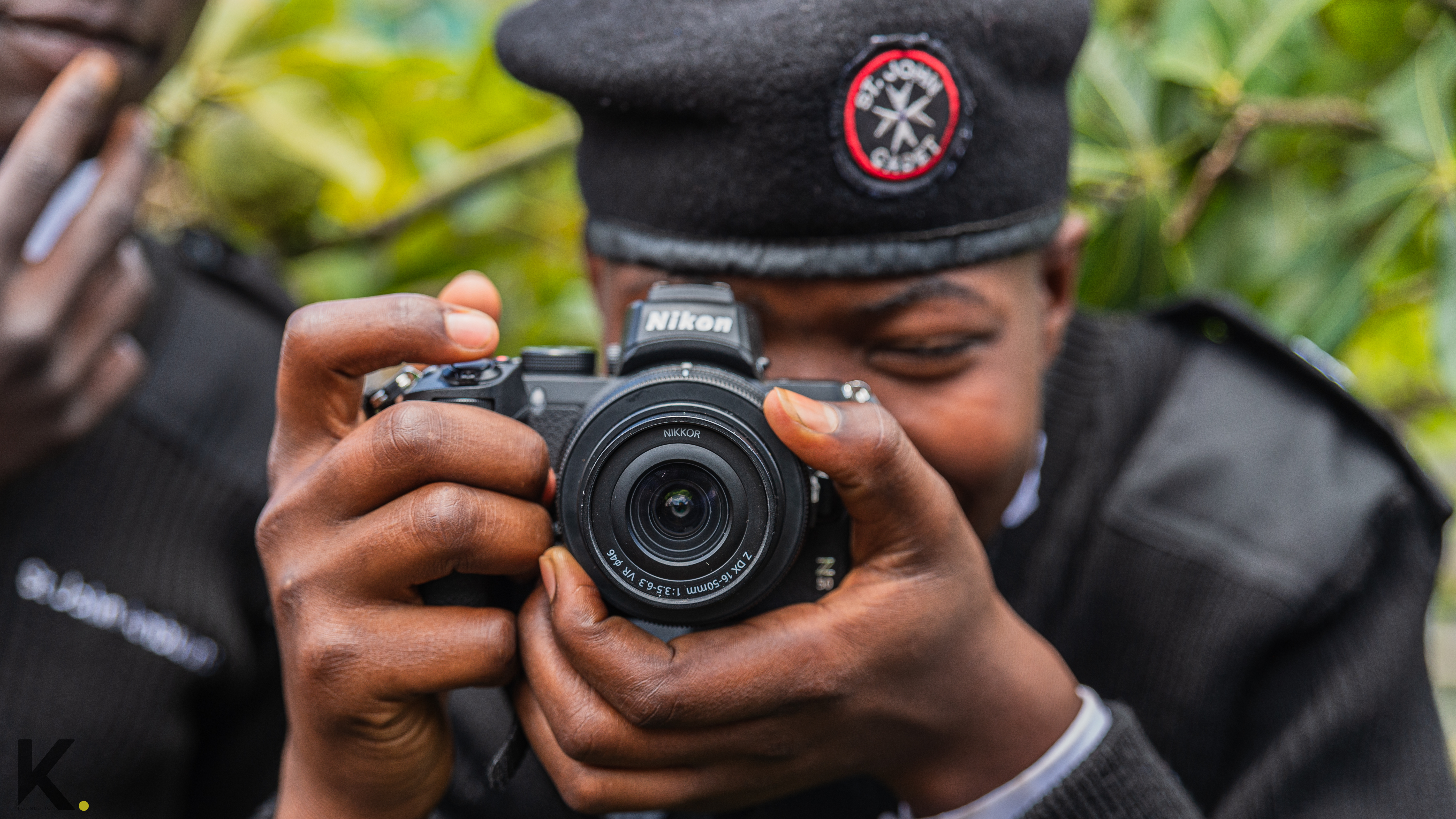 A person in a black cap holding a Nikon camera to their eye, smiling.